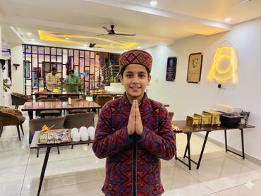 A boy in traditional attire greets with folded hands in a cozy dining area.