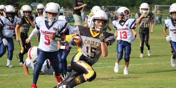 Youth football player from Chiefs team running with the ball, chased by Wolverines players.