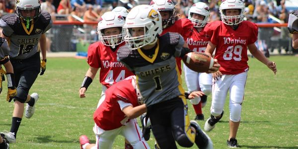 Youth football player in black and yellow runs with the ball past opponents in red.