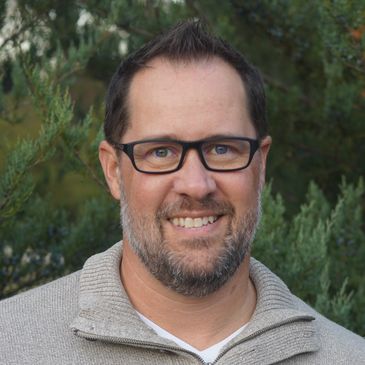 Smiling man with glasses in a beige sweater outdoors.