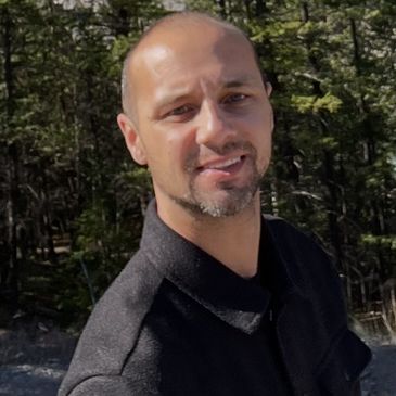 A man smiling outdoors with trees in the background.