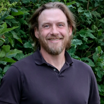 Smiling man with beard standing outdoors in front of green foliage.