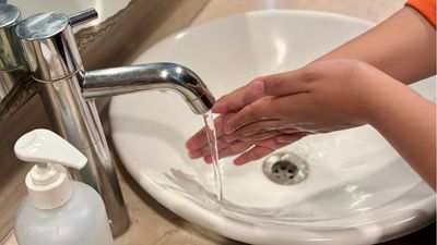 Person washing hands under a running faucet in a bathroom sink.