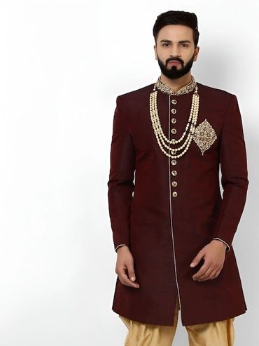 Man in traditional maroon sherwani with pearl necklace and gold pants.