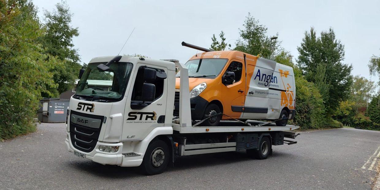 White tow truck carrying an orange and white van on a road surrounded by greenery with south coast recovery