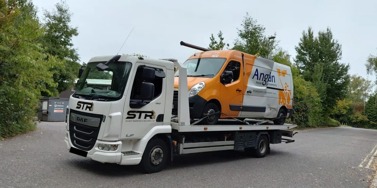 White tow truck carrying an orange and white van on a road surrounded by greenery with south coast recovery