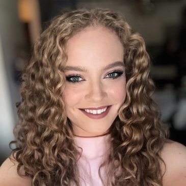 Smiling woman with curly hair and makeup in a softly lit room.