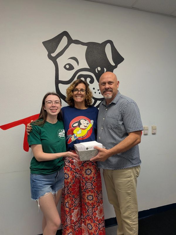 Three people smiling and holding a box, standing in front of a dog mural on the wall.