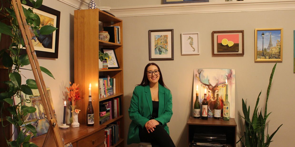 Tiana Desiree Grandilli in her art studio, seated and smiling before a gallery of her works.
