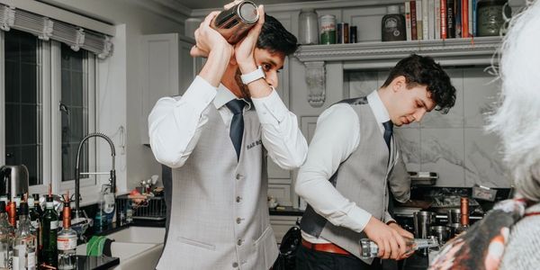 Two bartenders in gray vests preparing drinks in a kitchen.