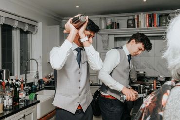 Two bartenders in gray vests preparing drinks in a kitchen.