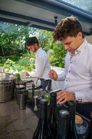 Two bartenders preparing drinks at an outdoor bar with greenery in the background.
