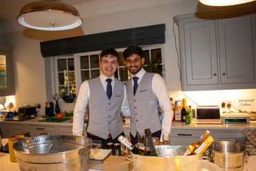 Two bartenders in matching vests smiling behind a counter with drinks.