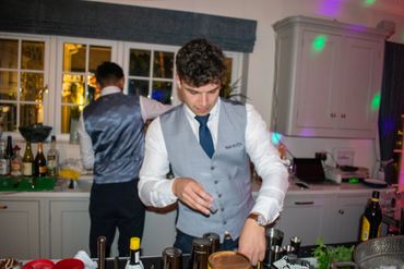 Two bartenders in vests preparing drinks behind a bar at night.
