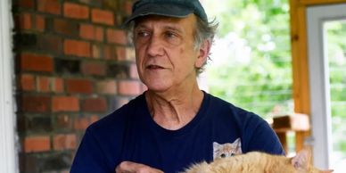 An older man holding a fluffy orange cat indoors near a brick wall.