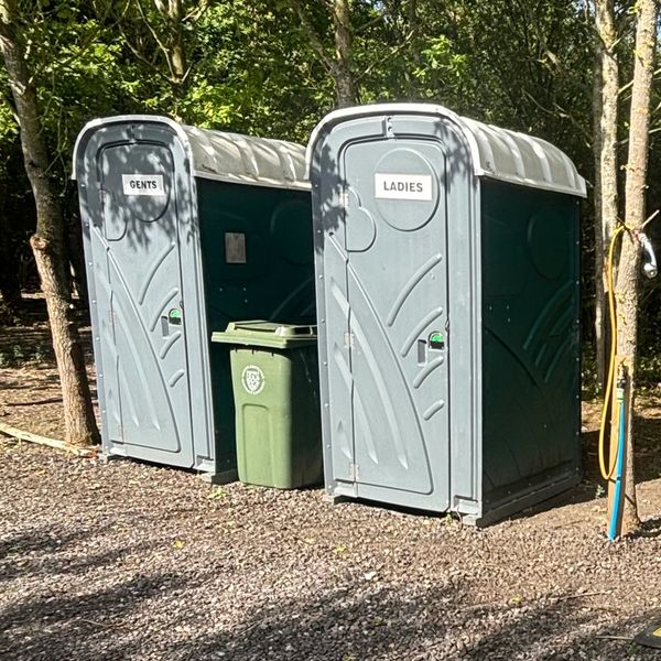 Two portable toilets labeled 'Gents' and 'Ladies' in a wooded outdoor area.