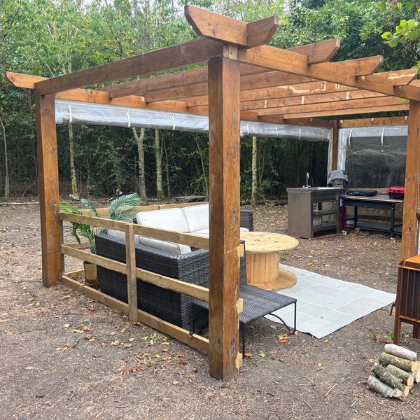 Outdoor wooden pergola with seating and a round table in a forest setting.