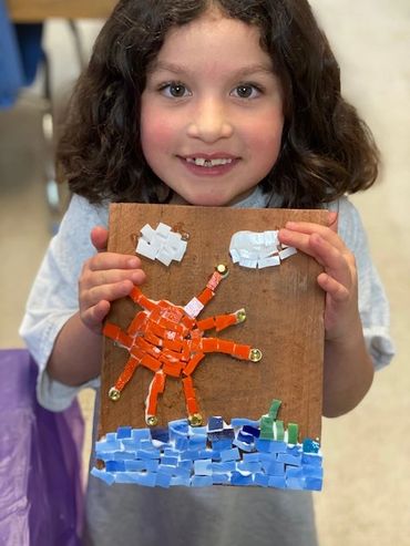 A child proudly holds a colorful mosaic art piece of a sun, clouds, and water.