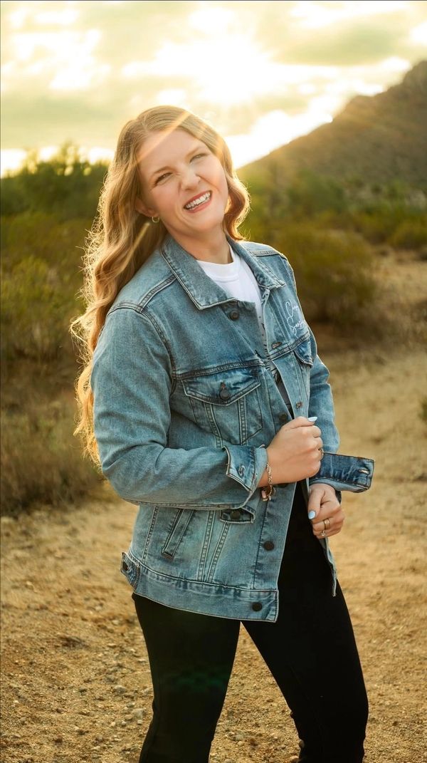 Smiling woman wearing a denim jacket in a sunny outdoor setting.