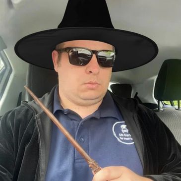 Man in sunglasses and a witch hat holding a wand inside a car.