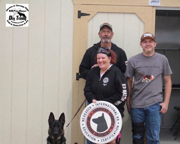 Three people with a trained dog in front of a training facility.