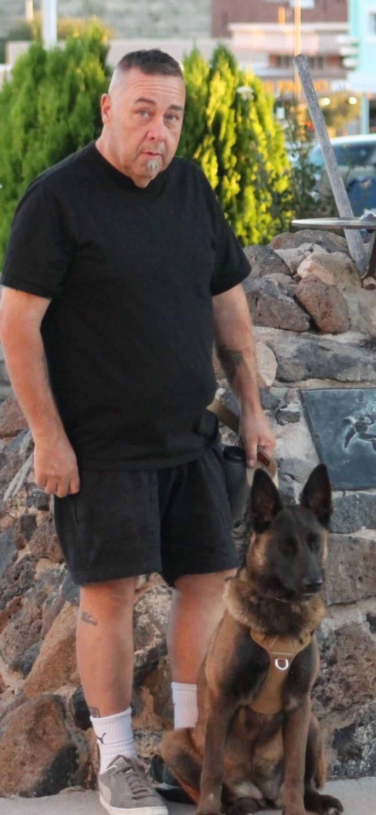 Man in black outfit standing next to a brown dog with a harness outdoors.