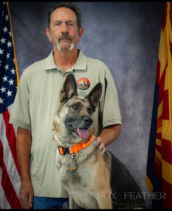 Man posing with a German Shepherd in front of American and Arizona flags.
