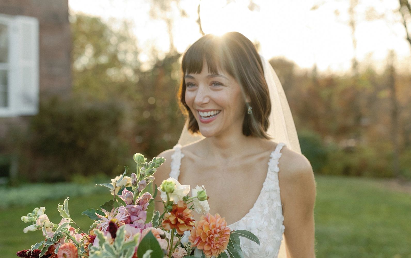 Smiling bride holding colorful bouquet outdoors in soft sunlight.