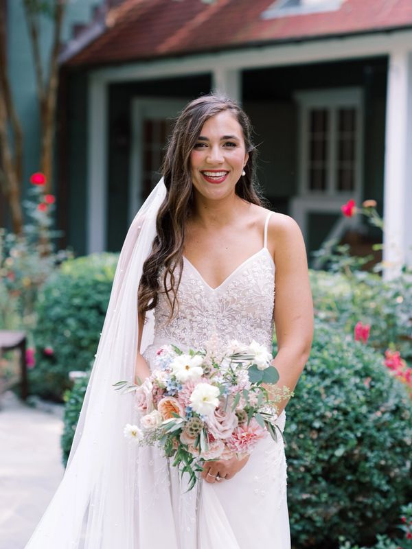 Smiling bride in a white dress holding a pastel flower bouquet outdoors.