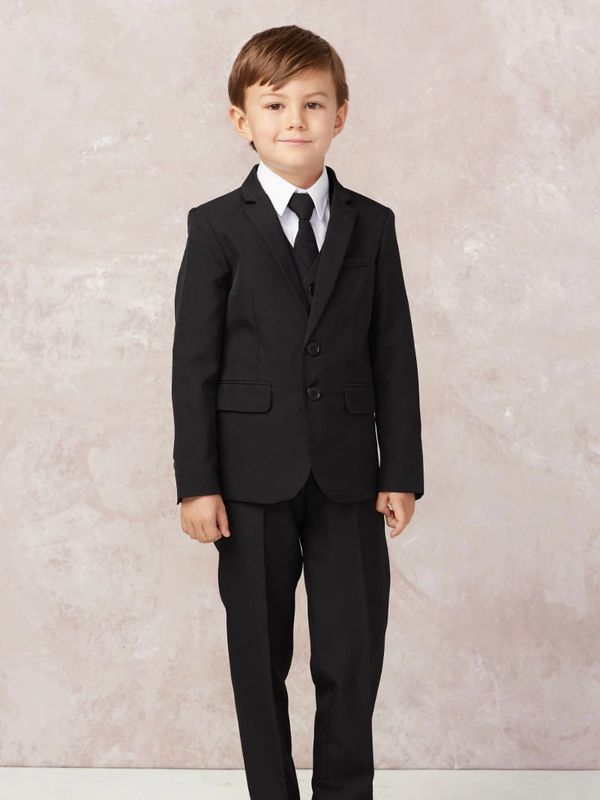 A young boy dressed in a formal black suit with a tie, standing confidently.