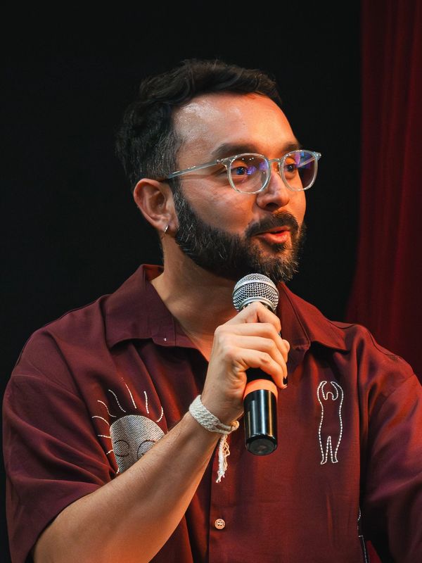 Man speaking into a microphone wearing glasses and a maroon shirt.