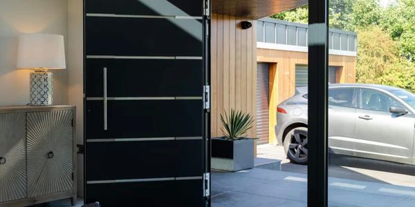 Modern black front door opens to driveway with silver car and greenery.