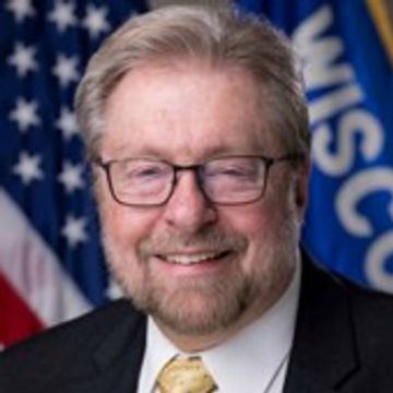 Smiling man with glasses in front of American and Wisconsin flags.