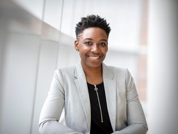 Black professional woman with short hair, grey jacket, black top in office building.