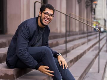 Young professional black man sitting on steps outside, with glasses, jacket and big smile.