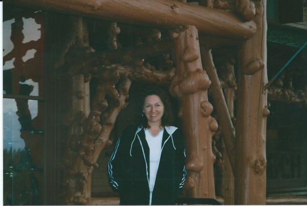 A woman smiling, standing by a rustic wooden structure with natural log details.