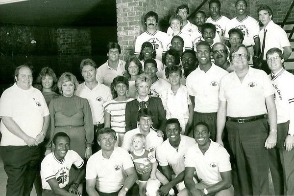 A large group photo of men and women, some in matching shirts, indoors by a brick wall.