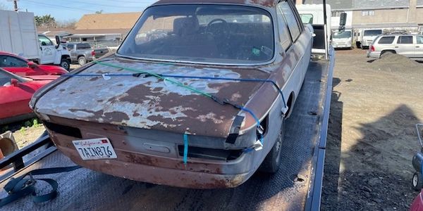 Rusty, damaged car secured on a flatbed truck in a lot.