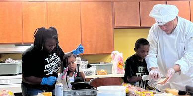 Adults and kids cooking together in a kitchen, wearing gloves and chef attire.