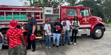 Children and a firefighter stand by a red fire truck on a sunny day.