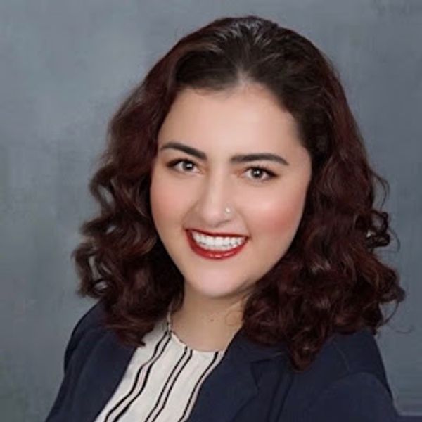 A smiling woman with curly dark hair wearing a navy blazer.