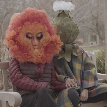 Two people in surreal, artistic costumes sitting on a bench.
