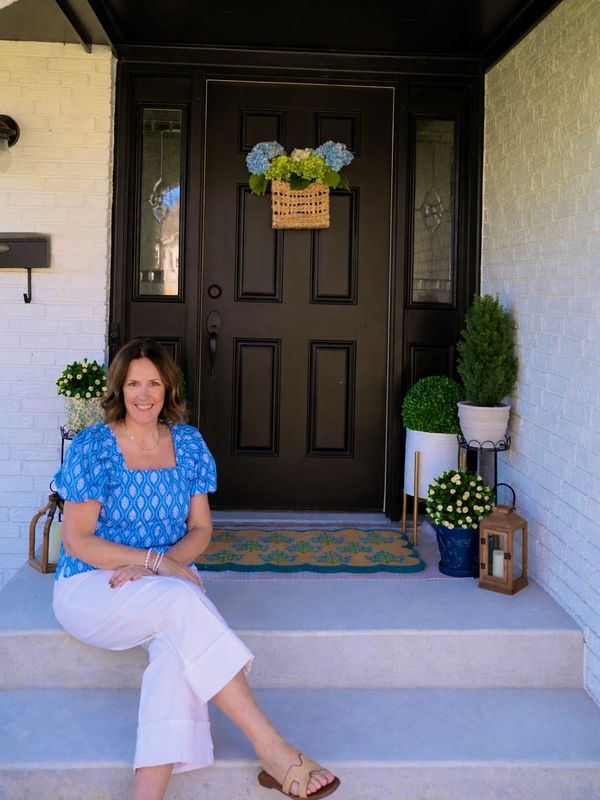 Woman in blue top sitting on porch steps in front of a black door with flower decor.