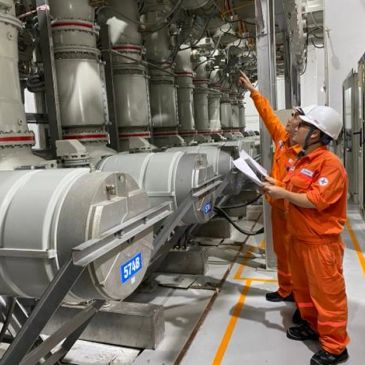 Two workers in orange uniforms inspecting industrial machinery.