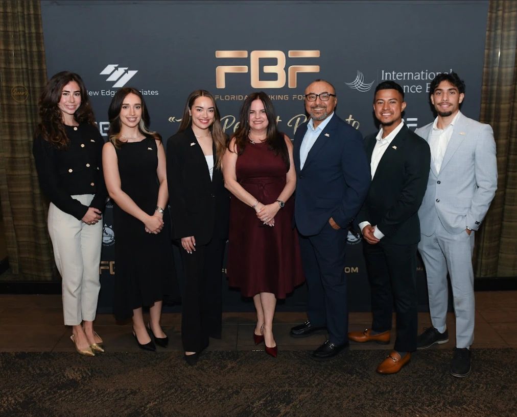 Seven professionally dressed people posing at a formal event with FBF backdrop.