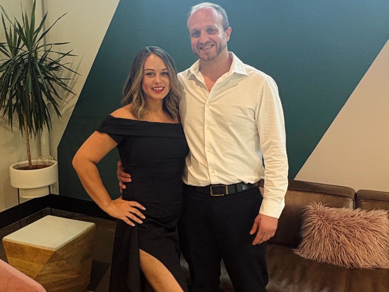 A couple dressed elegantly, smiling indoors near a geometric wall and stylish furniture.