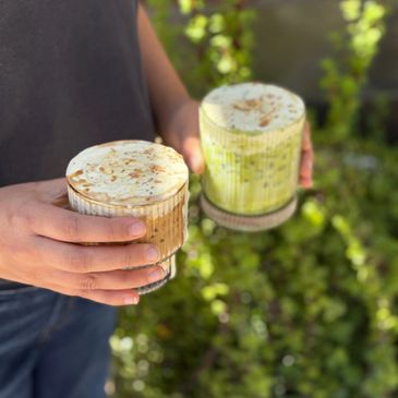 Person holding two layered beverages topped with foam outdoors.