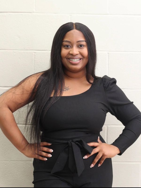Smiling woman in a stylish black outfit poses confidently against a light brick wall.