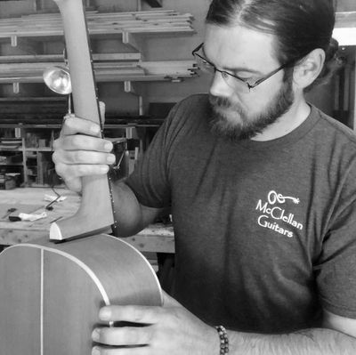 Luthier Matt McClellan, a man with glasses & dark hair in a ponytail, fits a guitar neck to the body