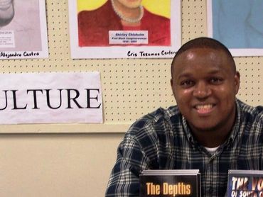 Smiling man at a book display with cultural posters in the background.